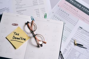 Post-it note with the words "tax deadline" next to a pair of glasses on top of a diary, with tax forms underneath